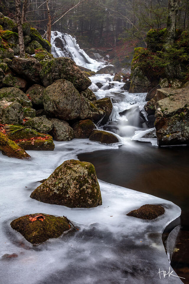 Limited edition print of a waterfall in the distance with the stream below half covered in ice that has enveloped three rocks in the water.